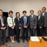 Seven business professionals in suits stand side by side in a conference room, posing for a formal group photo; several wear name badges on blue lanyards, with a table and documents visible on the right.