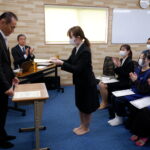 Woman in business attire hands a certificate to a barefoot recipient as masked coworkers applaud at an indoor ceremony.