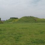 Green field with a grassy hill and a path up to the top, where a person stands.